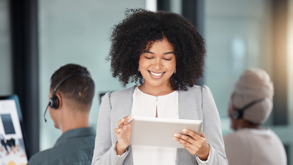 Smiling woman using a tablet with colleagues wearing headsets in the background, illustrating the use of CRM technology as part of balancing short term and long term marketing strategies. 