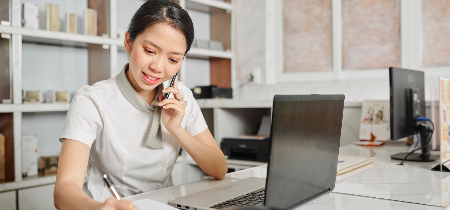 Resepsionis wanita Asia membuka laptopnya dan menggunakan ponsel cerdasnya secara bersamaan untuk mencatat booking langsung.